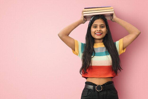 Sonriente joven estudiante india asiática posando libros de equilibrio aislados en su cabeza mirando directamente a la cámara mujer de cabello oscuro expresando emociones positivas