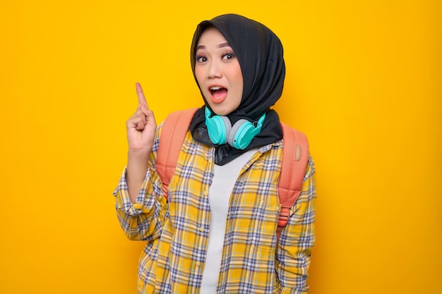 Sonriente joven estudiante musulmana asiática en camisa a cuadros con auriculares y mochila señalando con el dedo el espacio de copia aislado sobre fondo amarillo Concepto de colegio universitario de escuela de educación