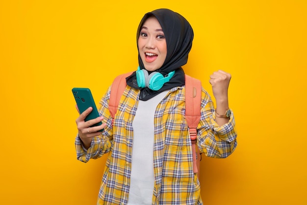 Sonriente joven estudiante musulmana asiática en camisa a cuadros con auriculares y mochila usando teléfono móvil y haciendo gesto de ganador sobre fondo amarillo Concepto de colegio universitario de escuela de educación