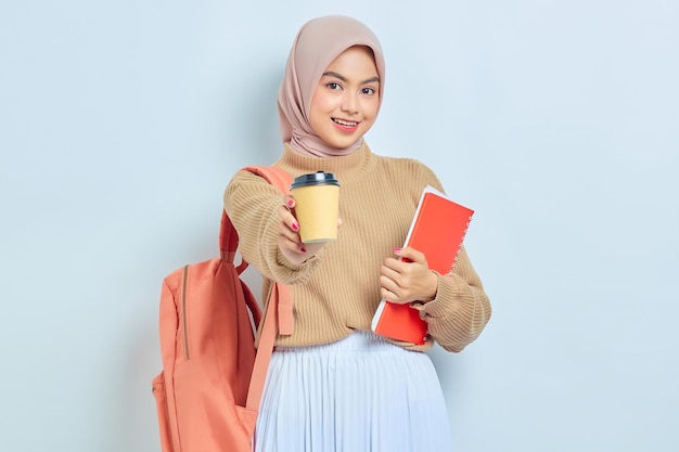 Sonriente joven estudiante musulmana asiática en suéter marrón con mochila sosteniendo libro y taza de café o té aislado sobre fondo blanco concepto de regreso a la escuela