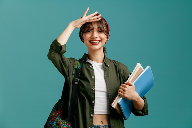 Sonriente joven estudiante con pañuelo y mochila sosteniendo blocs de notas con bolígrafo manteniendo la mano en la frente mirando a la cámara a distancia aislada en el fondo azul