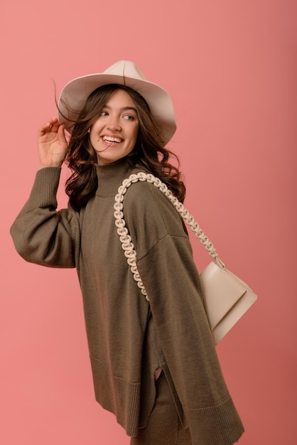 Sonriente joven europea con cabello oscuro que fluye en el sombrero mira hacia otro lado sobre fondo rosa Personas emociones estilo de vida y concepto de moda