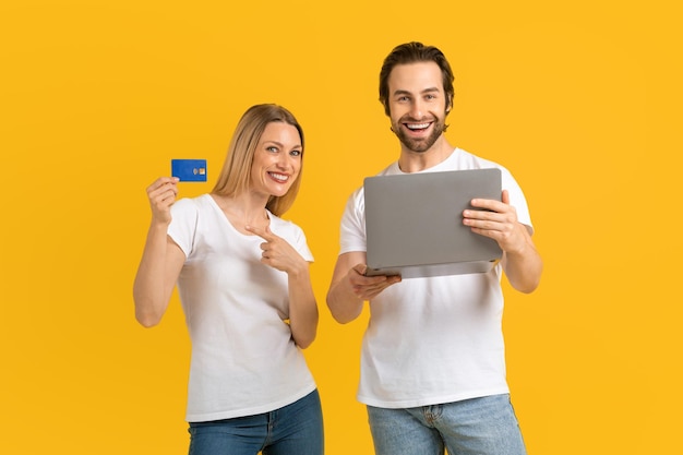 Sonriente joven europea y hombre con camisetas blancas miran la computadora portátil muestran la tarjeta de crédito