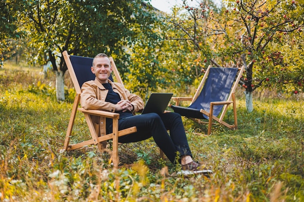 Sonriente joven freelancer sentado en una silla de madera con una computadora portátil mientras trabaja al aire libre en el jardín Trabajo remoto
