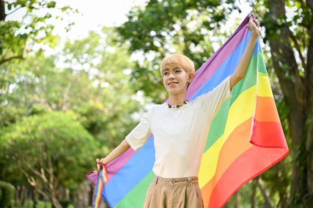 Sonriente joven gay asiático con una bandera del arco iris LGBT de pie en el parque verde