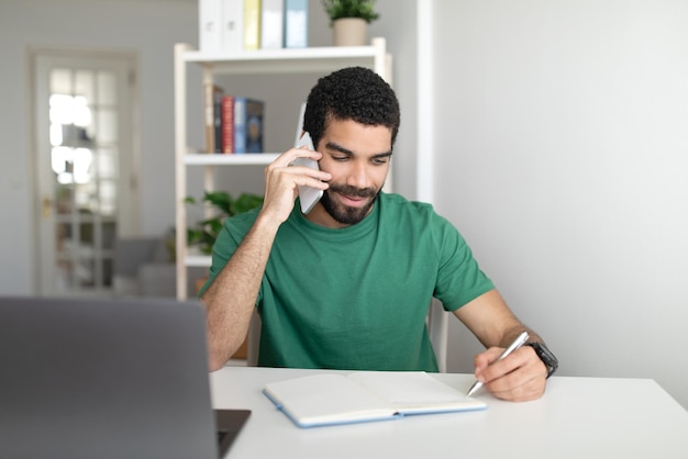 Sonriente joven gerente de medio oriente en la mesa con una computadora portátil que trabaja de forma remota llamada por teléfono inteligente