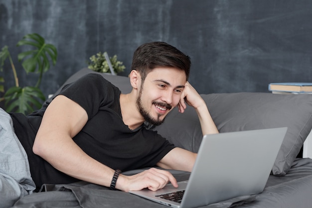 Sonriente joven guapo acostado en la cama y usando la computadora portátil mientras sombrea con amigos la aplicación en línea