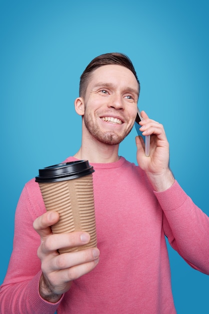 Sonriente joven guapo con rastrojo tomando café de la taza para llevar y charlando por teléfono celular