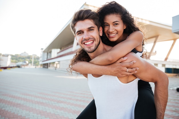 Sonriente joven guapo sosteniendo a su novia en la espalda al aire libre