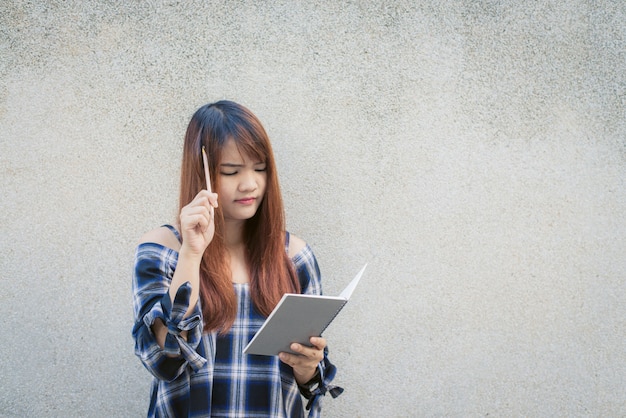 Sonriente joven hermosa mujer asiática pensando con libro de escritura sobre fondo de muro de hormigón