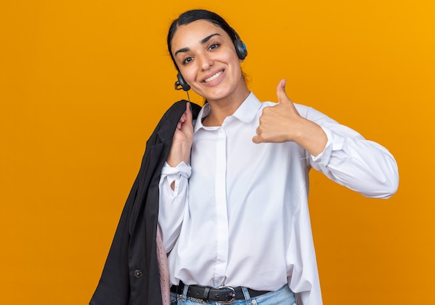 Sonriente joven hermosa mujer con auriculares poniendo la chaqueta en el hombro mostrando el pulgar hacia arriba