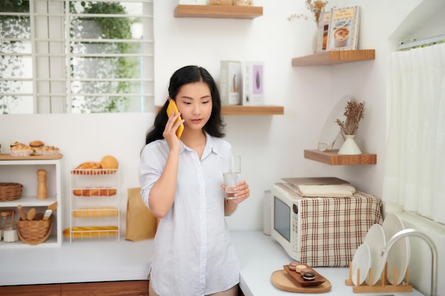 Sonriente joven hermosa mujer de pie bebiendo agua y hablando por teléfono inteligente en la cocina