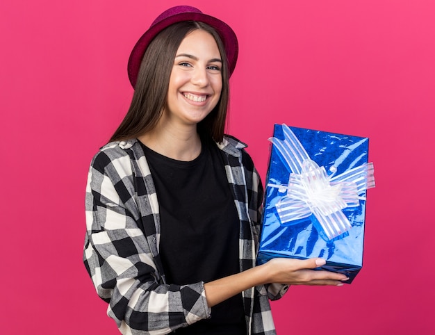 Sonriente joven hermosa mujer vistiendo gorro de fiesta con caja de regalo aislada en la pared rosa