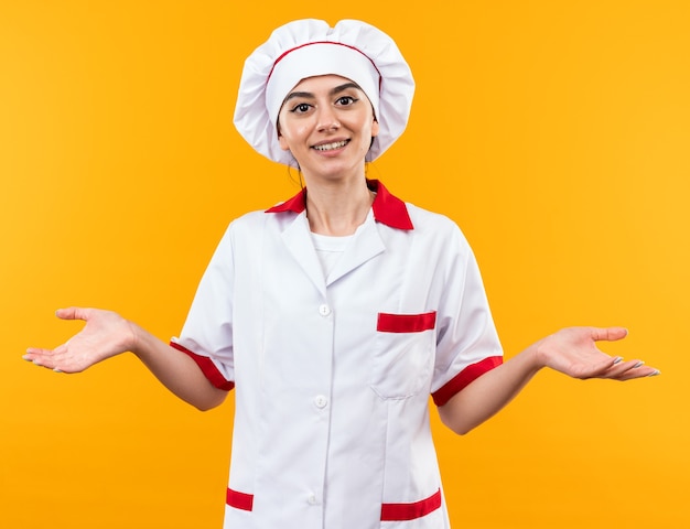 Sonriente joven hermosa en uniforme de chef cruzando las manos aisladas en la pared naranja