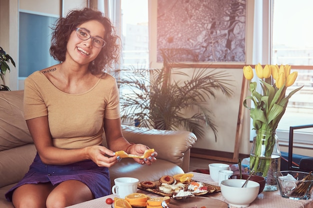 Sonriente joven hispana rizada desayunando en casa sentada en un sofá.