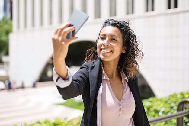 Sonriente joven hispanoamericana tomando un selfie al aire libre con su teléfono inteligente. Espacio para texto.