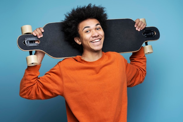 Sonriente joven hombre afroamericano sosteniendo longboard aislado sobre fondo azul.