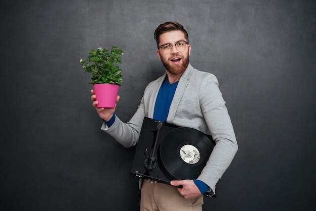 Sonriente joven hombre de negocios barbudo con gafas sosteniendo flores en maceta y tocadiscos