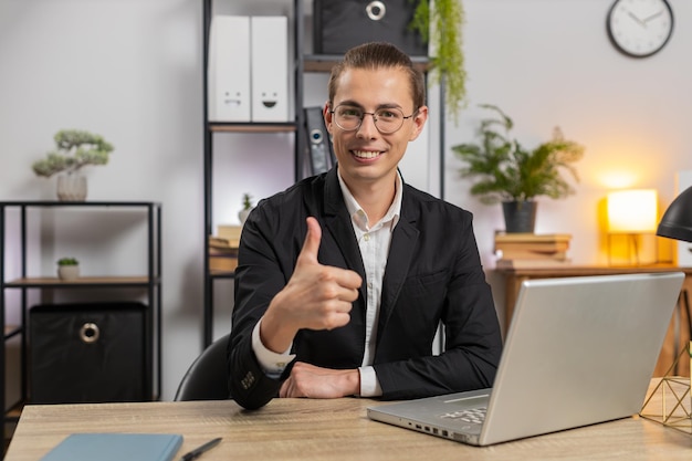 Sonriente joven hombre de negocios caucásico freelance mostrando pulgares dobles sentado en el escritorio de la oficina de casa