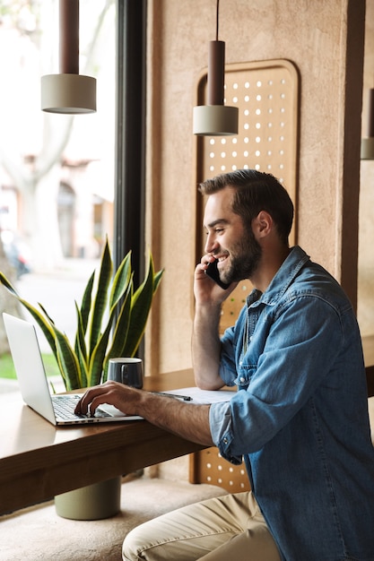 Sonriente joven hombre vestido con camisa de mezclilla hablando por teléfono móvil con portátil y portapapeles mientras trabajaba en el café en el interior