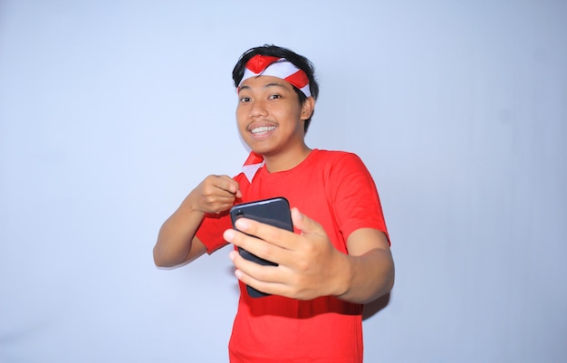 Sonriente joven indonesio apuntando al teléfono móvil con cara feliz y emocionada con camiseta roja