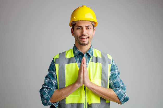 Sonriente joven ingeniero masculino con casco de seguridad y uniforme mirando a la cámara mostrando un gesto de namaste aislado en fondo blanco