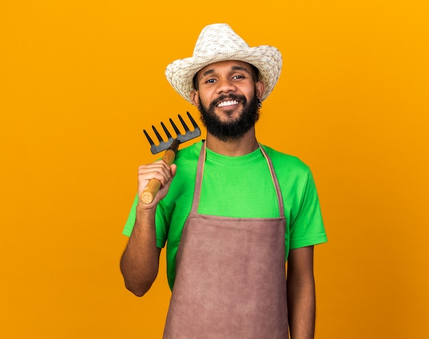Sonriente joven jardinero afroamericano vistiendo sombrero de jardinería sosteniendo el rastrillo en el hombro