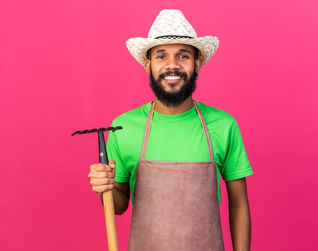 Sonriente joven jardinero afroamericano vistiendo sombrero de jardinería sosteniendo rastrillo