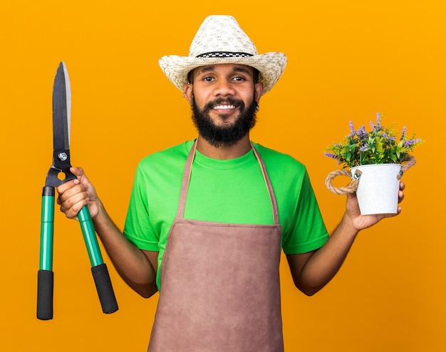 Sonriente joven jardinero afroamericano vistiendo sombrero de jardinería sosteniendo tijeras y flor en maceta