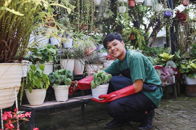 Sonriente joven jardinero asiático en delantal sosteniendo planta en maceta en el invernadero