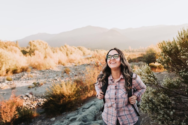 Foto sonriente joven latina feliz con gafas y una mochila, caminando en un hermoso paisaje.