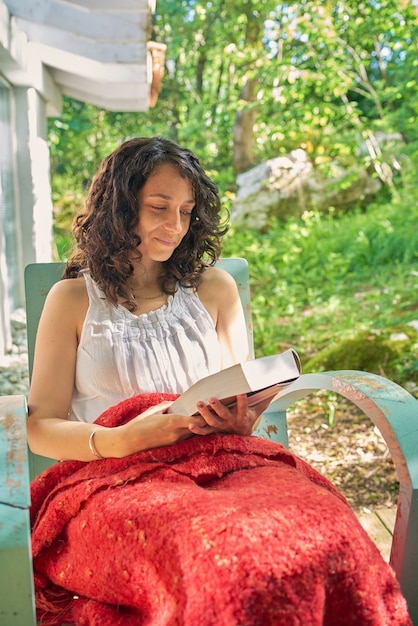 Sonriente joven leyendo un libro sentada en su porche se sienta en una silla vintage turquesa