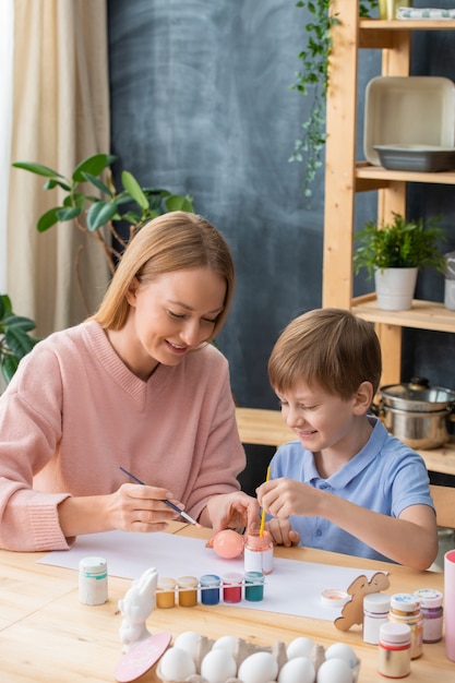 Sonriente joven madre sentada a la mesa y pintando huevos de Pascua junto con lindo hijo