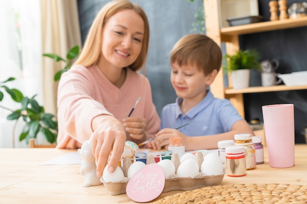 Sonriente joven madre y su hijo sentados a la mesa y disfrutando de pintar huevos de Pascua