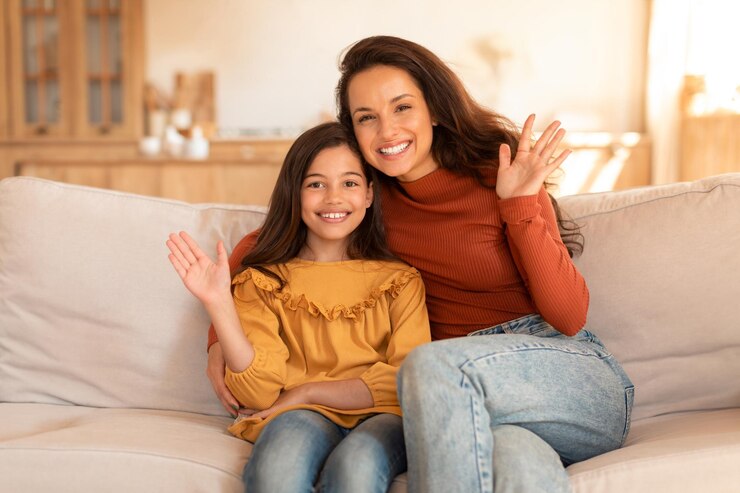 Sonriente joven mamá y su hija saludando con la mano en casa
