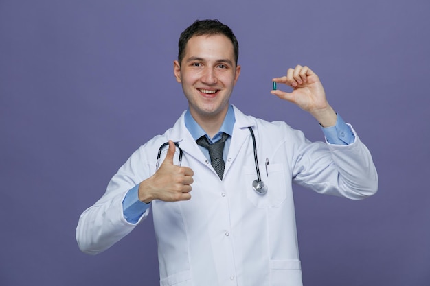 Sonriente joven médico masculino con bata médica y estetoscopio alrededor del cuello mostrando cápsula mirando a la cámara mostrando el pulgar hacia arriba aislado en fondo púrpura