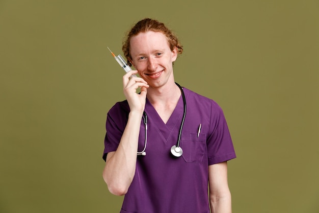 Sonriente joven médico masculino vistiendo uniforme con estetoscopio sosteniendo jeringa aislado sobre fondo verde