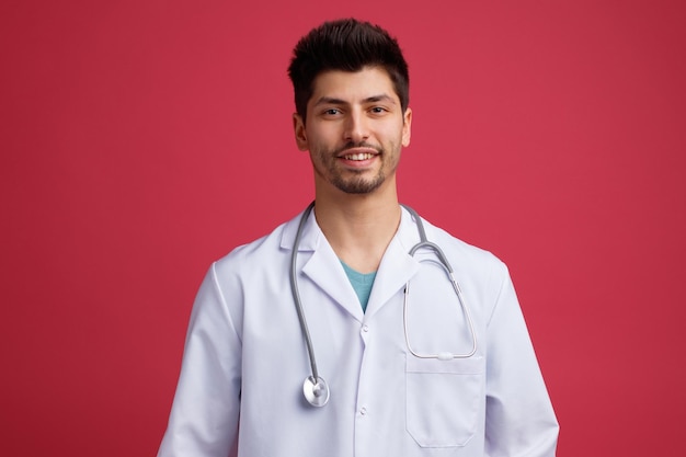 Sonriente joven médico masculino vistiendo uniforme médico y estetoscopio alrededor de su cuello mirando a la cámara aislada sobre fondo rojo.