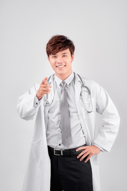Sonriente joven médico positivo en bata de laboratorio apuntando hacia afuera, aislado en blanco