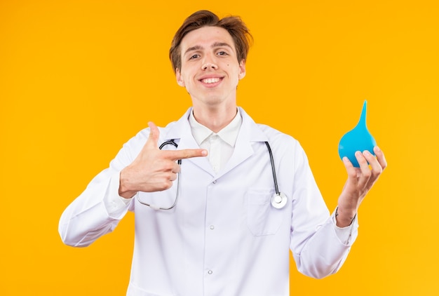 Sonriente joven médico vistiendo bata médica con estetoscopio sosteniendo y puntos en el enema aislado en la pared naranja