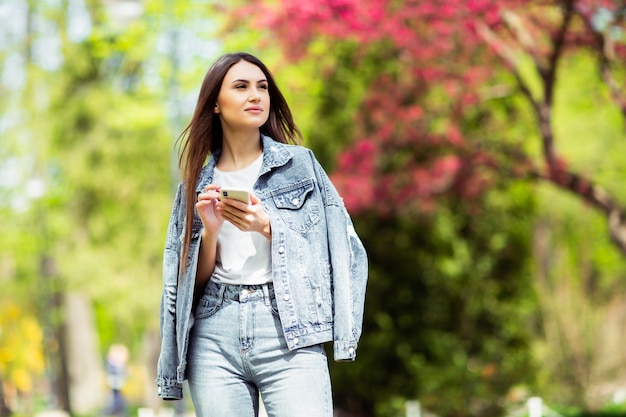 Sonriente joven morena caucásica enviando mensaje en el teléfono inteligente fuera solo Use ropa de jeans .Tecnologías y gadgets. Publicidad para tienda de venta de electricidad y teléfonos móviles.