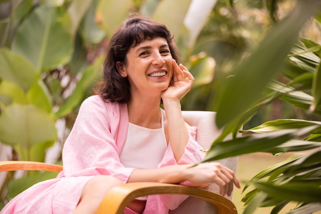 Sonriente joven morena caucásica vestida de rosa mira a la cámara sentada en una silla entre la naturaleza en el jardín Felicidad vacacional y concepto de placer