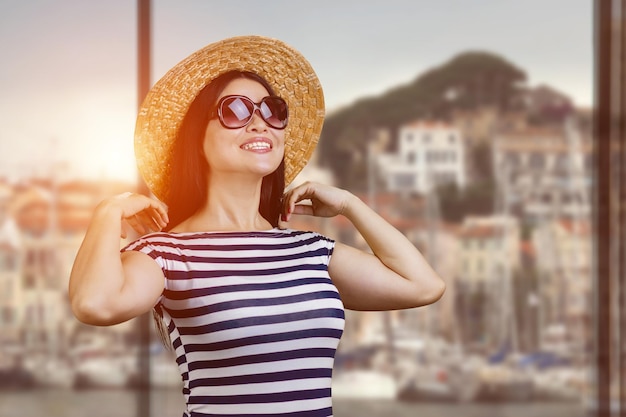 Sonriente joven morena con gafas de sol y paisaje urbano de sombrero de paja con puesta de sol en el fondo