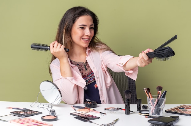 Sonriente joven morena sentada a la mesa con herramientas de maquillaje sosteniendo peines