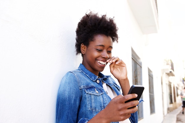Sonriente joven mujer africana apoyada contra la pared usando teléfono móvil