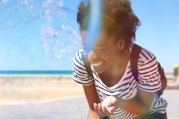 Sonriente joven mujer afro jugando con pompas de jabón al aire libre en la playa