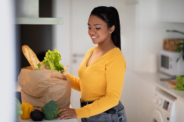 Sonriente joven mujer afroamericana en casual desempaqueta una bolsa de papel con comestibles y verduras orgánicas