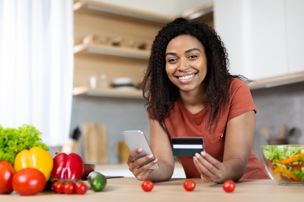 Sonriente joven mujer afroamericana con teléfono inteligente y compras con tarjeta de crédito en línea