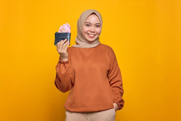 Sonriente joven mujer asiática en camisa naranja mostrando billetera llena de billetes de dinero aislado sobre fondo amarillo