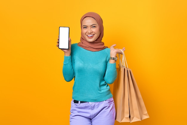 Sonriente joven mujer asiática mostrando la pantalla del teléfono inteligente y sosteniendo bolsas de la compra sobre fondo amarillo